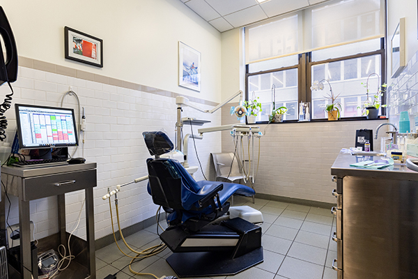 The image shows a dental office interior, featuring a dental chair with a patient s headrest and an adjustable dental lamp. A computer monitor is visible on the countertop next to the chair, displaying what appears to be a digital X-ray or dental imaging software. The room has a modern design with white walls, a tiled floor, and a window covered with blinds.