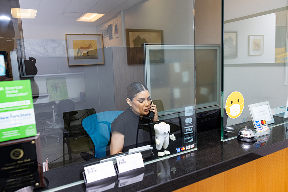 A woman is sitting at a reception desk in an office setting, smiling and looking towards the camera.
