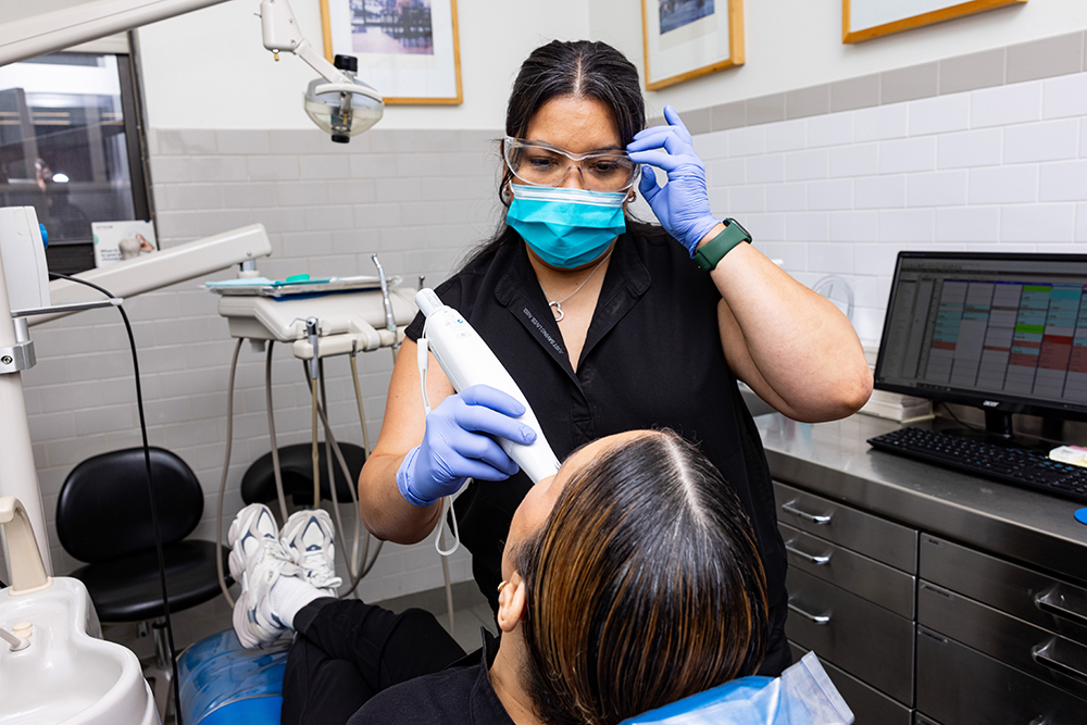 A dental hygienist in a black apron and face mask, using a dental drill on a patient s teeth, with a computer monitor displaying dental software in the background.