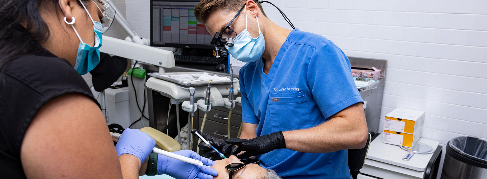 A person in a blue surgical gown and mask is performing dental work on another individual, who appears to be seated with their mouth open.