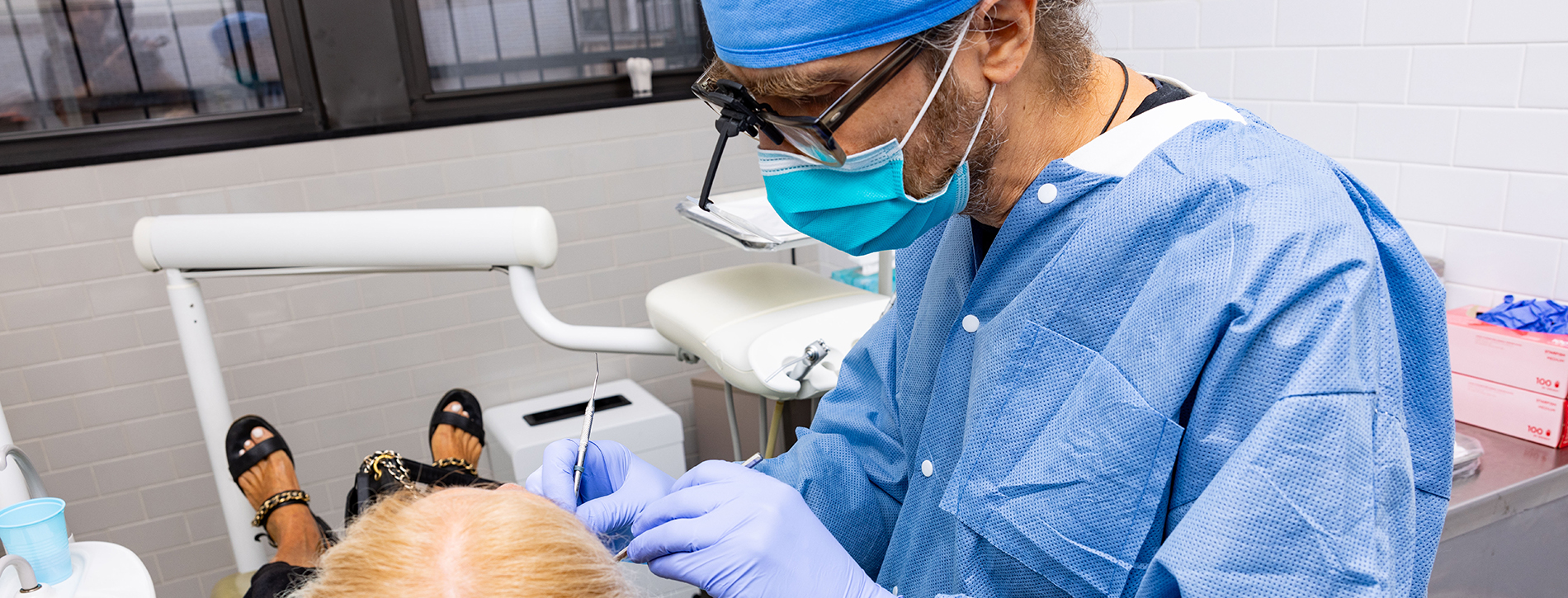 The image shows a dental professional in a surgical mask and gown, performing a procedure on a patient s mouth while seated in a dental chair.