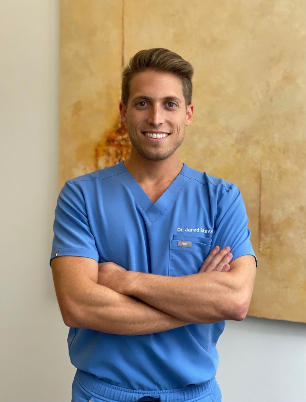 The image features a man in a blue scrub suit, standing with his arms crossed in front of him. He is smiling and appears to be posing for the photograph.