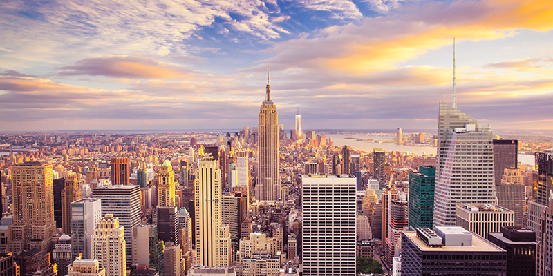 The image features a vibrant city skyline with the Empire State Building prominently visible, set against a backdrop of a clear blue sky with a few clouds.