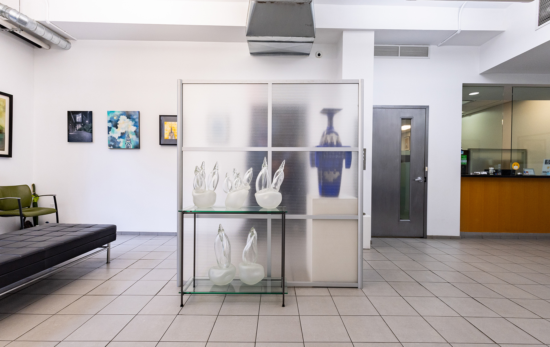 The image shows an interior space, likely a lobby or waiting area, with a display case containing various items, including what appears to be sculptural objects and possibly a vase. There is a bench in the foreground, and the flooring suggests a modern design. The room has a clean and minimalistic aesthetic.