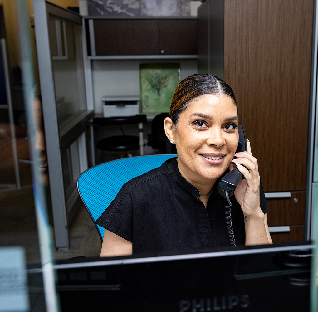 The image shows a woman sitting at a desk in an office, talking on the phone with a smile.