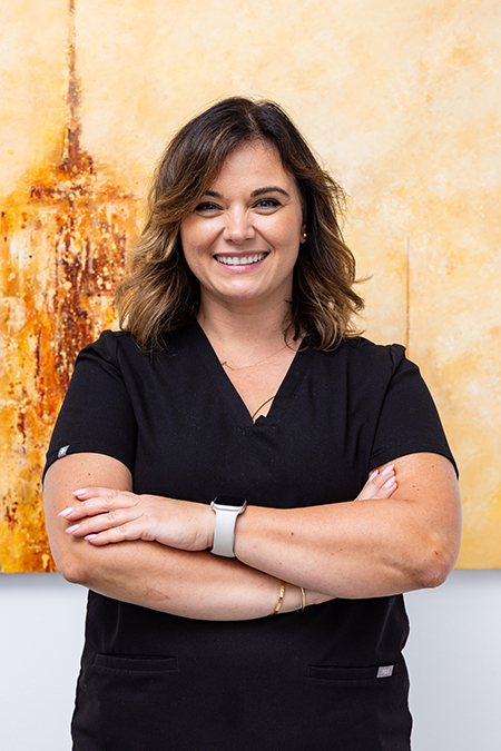 The image shows a woman standing in front of a painting, smiling at the camera. She is wearing a black shirt and has her arms crossed.