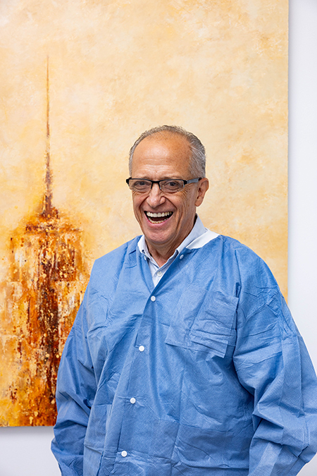 The image shows a man in a white lab coat standing in front of a painting, smiling and looking directly at the camera.