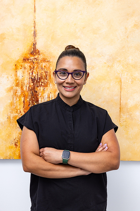 A woman in a black shirt and blue glasses stands confidently with her arms crossed, smiling slightly. She is positioned in front of an abstract painting featuring a cityscape.
