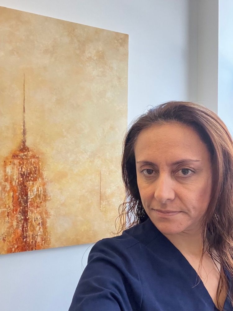 A woman stands in front of a framed artwork, posing for the camera with a neutral expression.