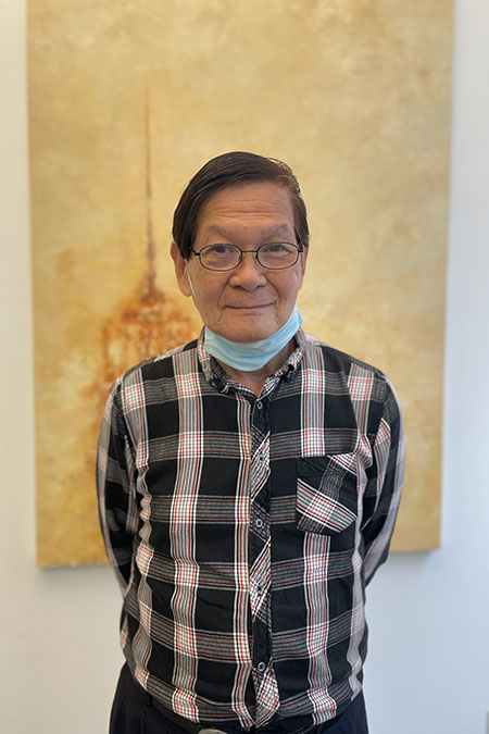 An elderly man in glasses stands against a wall, posing with a neutral expression and wearing a plaid shirt.