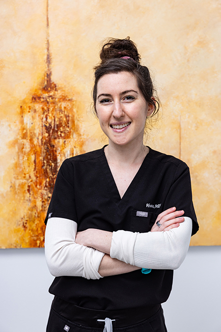 The image shows a woman standing in front of a large canvas, posing with her arms crossed. She is wearing scrubs and has short hair.