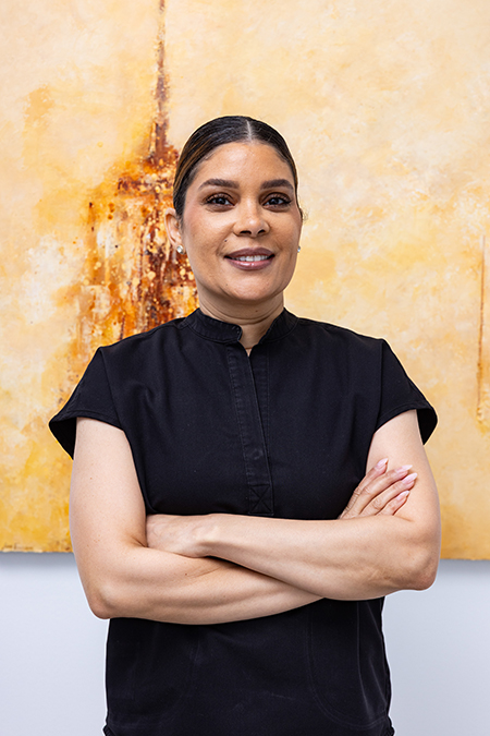 The image shows a woman standing in front of an abstract painting, wearing a dark top and black pants, with her arms crossed.