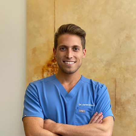 The image features a person who appears to be a male healthcare professional, likely a doctor or surgeon, standing in front of an interior wall with artwork and smiling at the camera. He is wearing a light blue shirt and dark pants, and there s a name tag visible on his attire that reads  Dr. Jared Styler.  The background has neutral tones and a warm lighting ambiance.