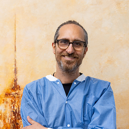 The image shows a man wearing glasses, a blue apron, and a white lab coat standing in front of a painting. He is smiling and has his arms crossed.