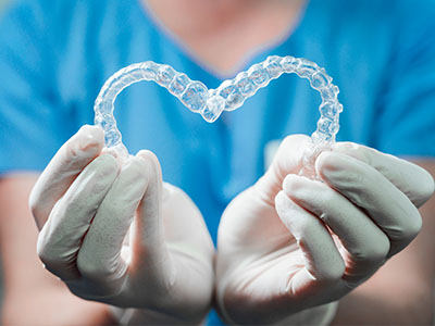 A person wearing a blue medical uniform and white gloves is holding a transparent plastic heart-shaped object that resembles dental retainers.