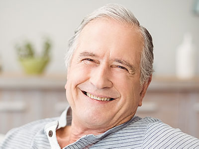 The image shows an elderly man with white hair, wearing a blue shirt and sitting in a relaxed pose. He is smiling and appears to be at ease.