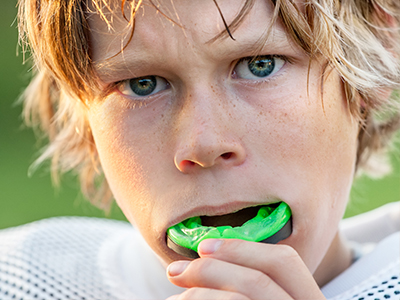 The image shows a young male with blonde hair and blue eyes, wearing a white football jersey. He is holding a green toothbrush in his mouth, which appears to be covered with toothpaste foam.